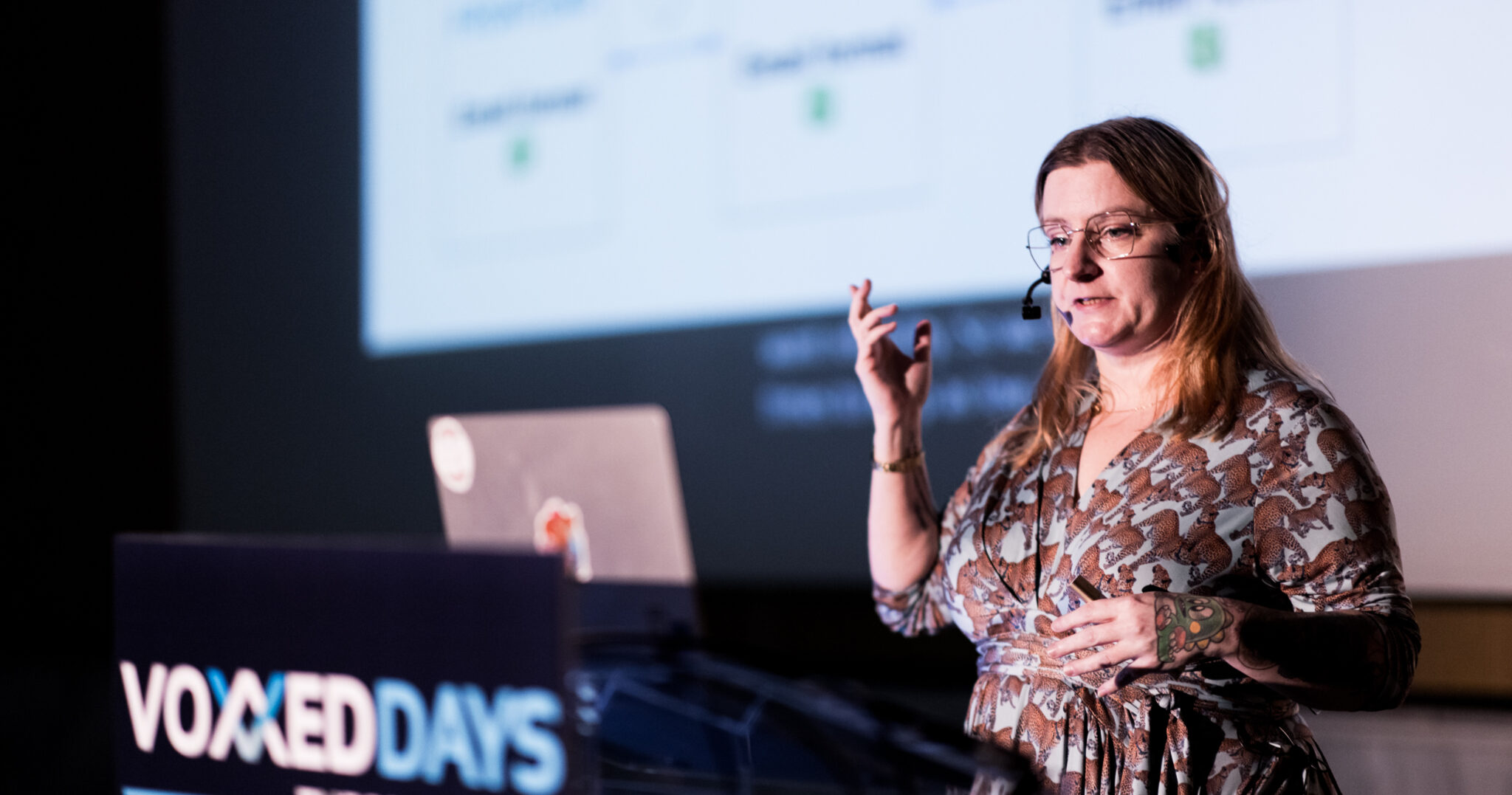 Picture of blonde woman in a patterned dress speaking on a stage at Voxxed Days Thessaloniki